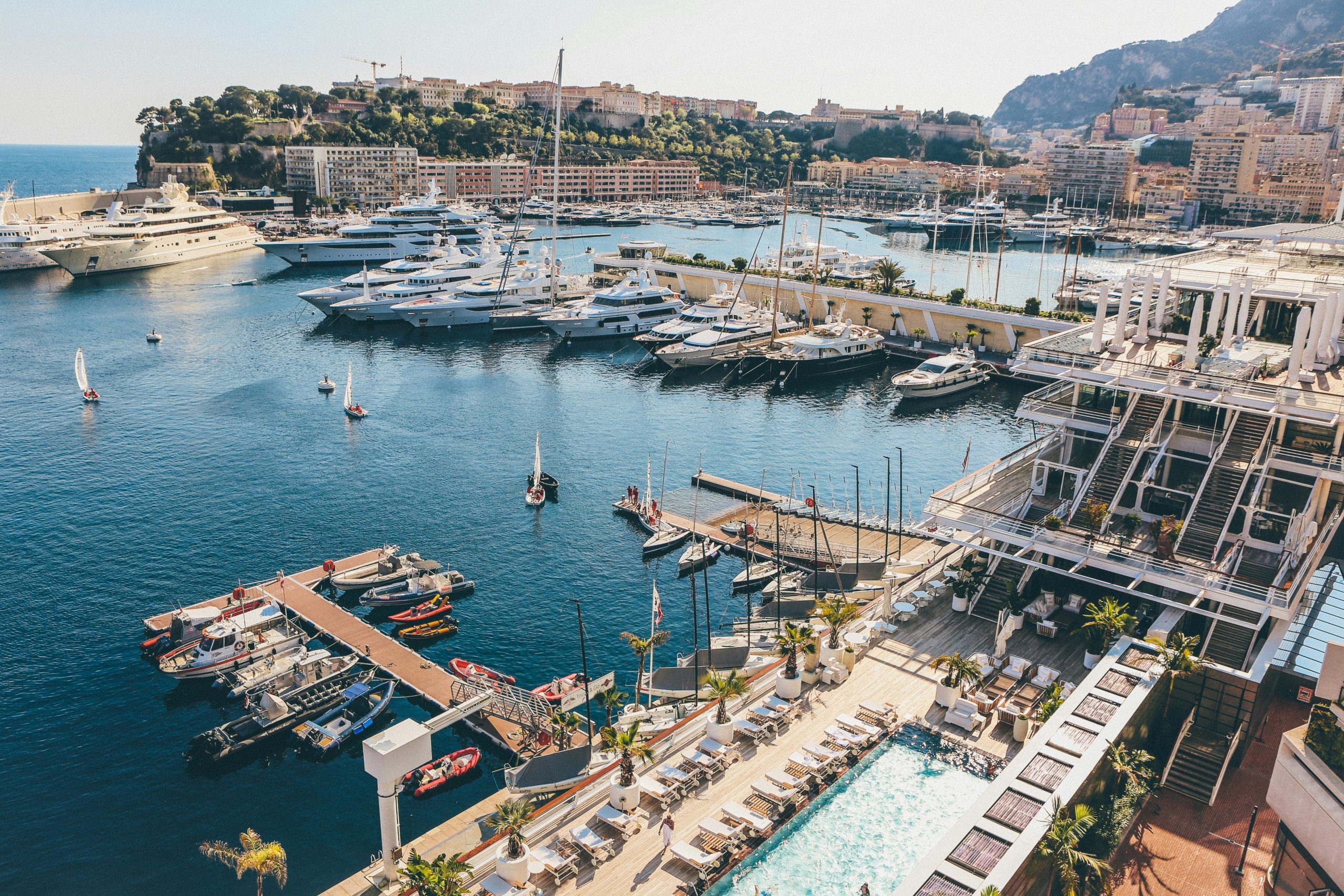 Une vue aérienne et en plongée du Port Hercule à Monaco sous un ciel radieux. Les eaux bleu azur sont parsemées de superyachts de luxe, de bateaux à moteur et de petits voiliers. Au premier plan, un bâtiment moderne à plusieurs niveaux dévoile une terrasse extérieure avec piscine, chaises longues et palmiers. En arrière-plan, le paysage urbain dense de Monaco s'élève à flanc de colline, mettant en valeur l'immobilier résidentiel haut de gamme surplombant la mer Méditerranée.